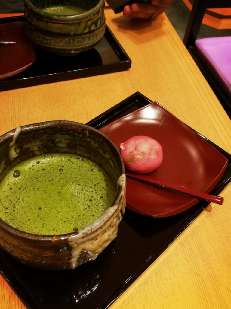 Matcha served in a ceramic chawan bowl with a small pink wagashi on a plate.