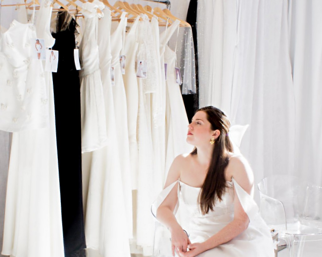 Woman sitting in front of a rack of wedding dresses and looking into the distance.