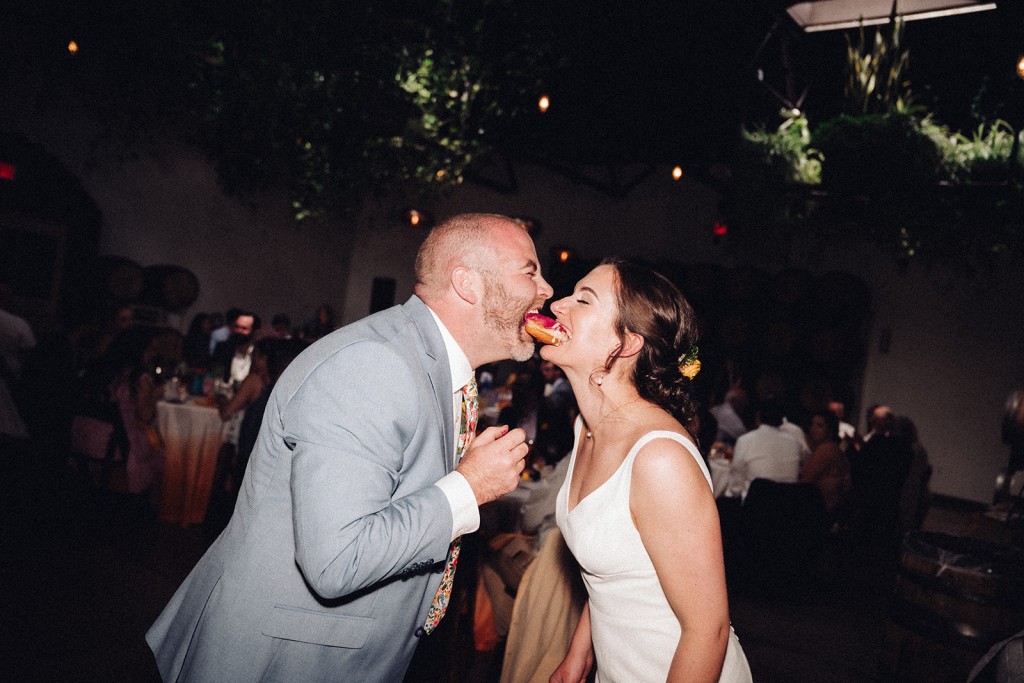 The newlyweds holding a shared donut by the mouth