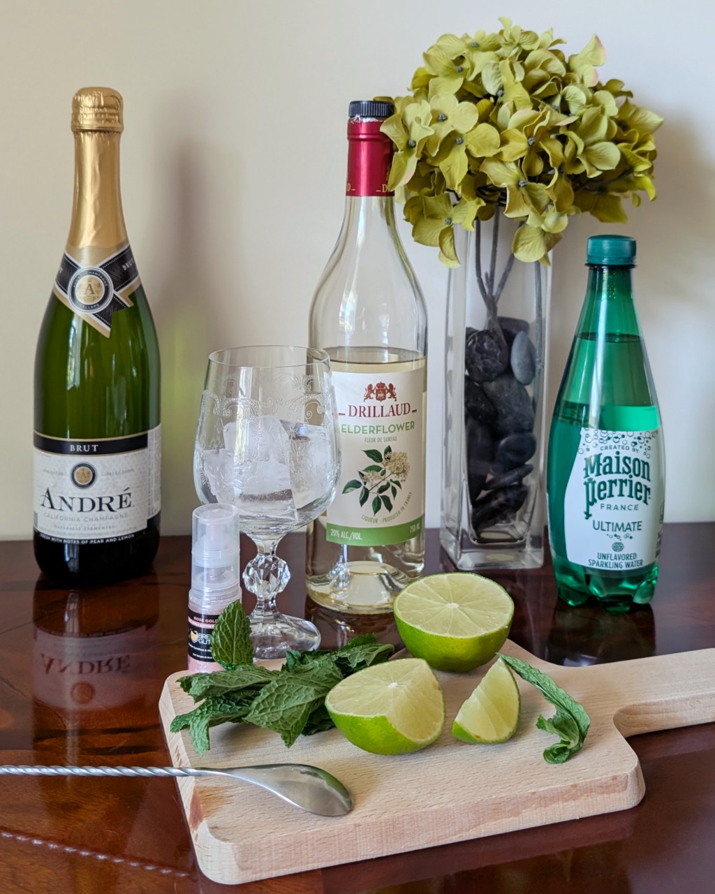 Ingredients assembled for a Sparkling Spritz cocktail with brut, elderflower liqueur, lime, mint, sparkling water, and edible glitter.